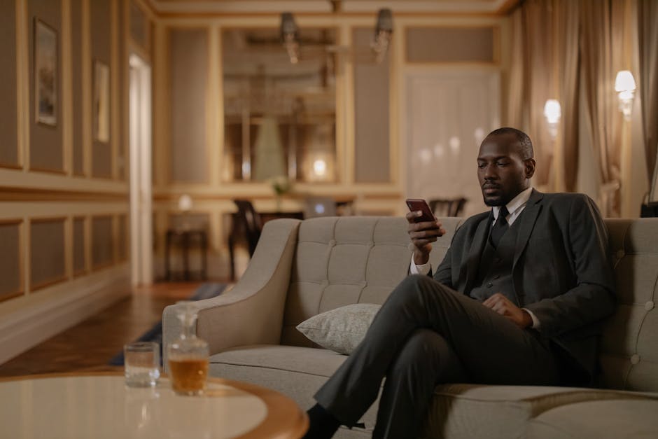 Elegant businessman using smartphone while relaxing in hotel lounge with whiskey on the table.