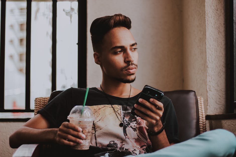Contemplative young man sitting indoors, holding a smartphone and drink, wearing casual attire.