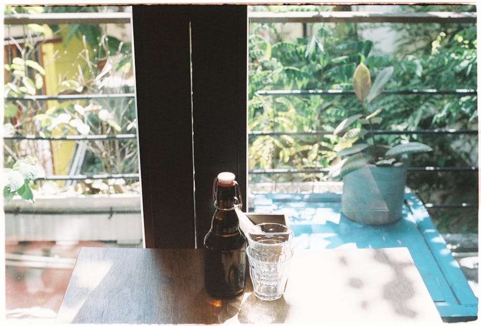 A bottle and glass on a sunlit table by a window with plants and balcony view.
