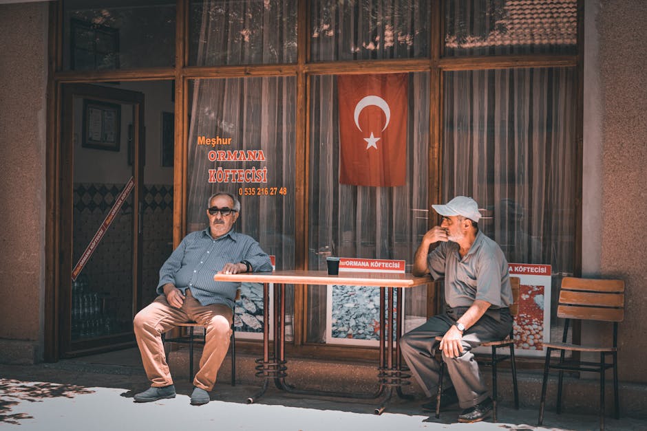 Two middle-aged men sitting outside a Turkish cafe adorned with a national flag.