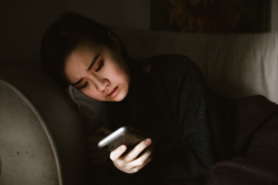 A young woman lying on a couch looking at her smartphone in a dimly lit room.