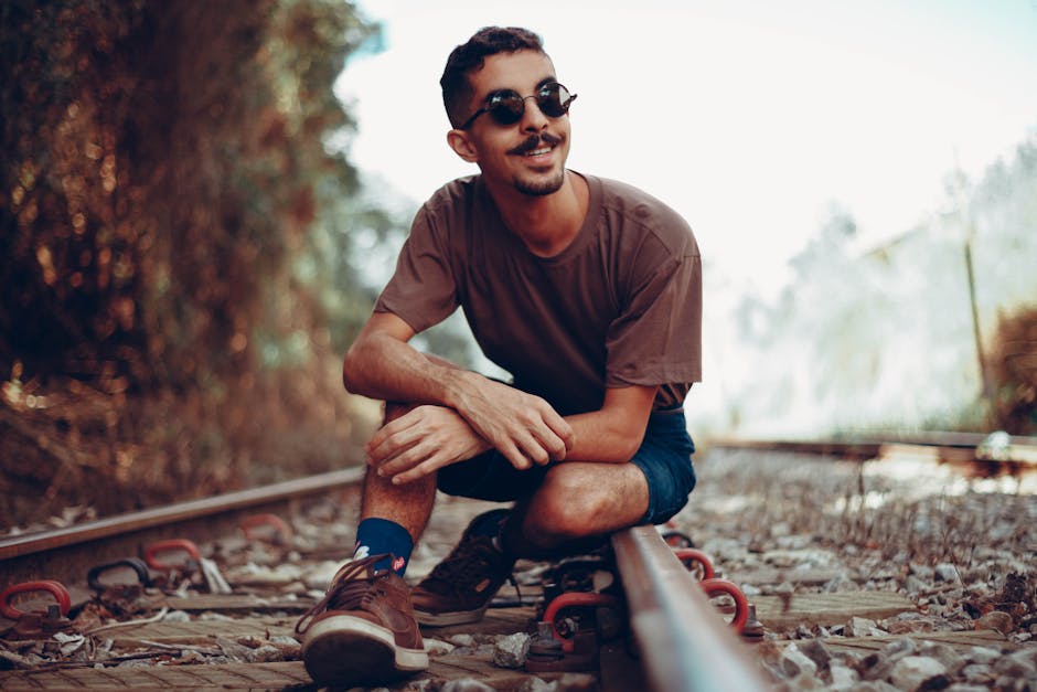 Fashionable man in sunglasses sitting on railway tracks, smiling on a sunny day.