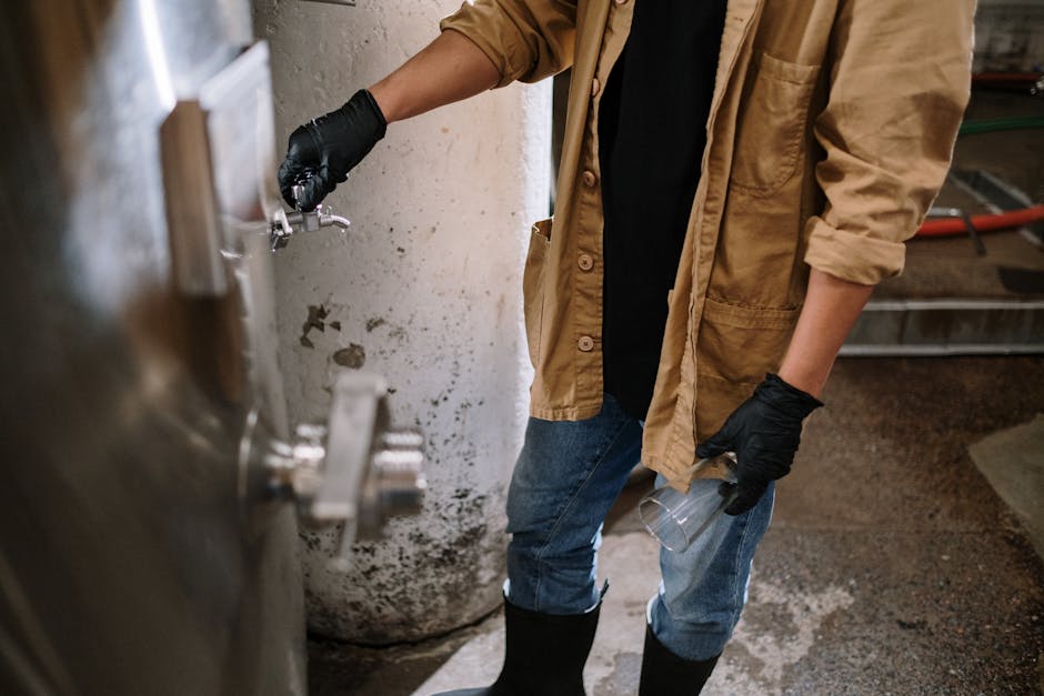A brewmaster wearing gloves operates a beer tap in an industrial brewery setting.