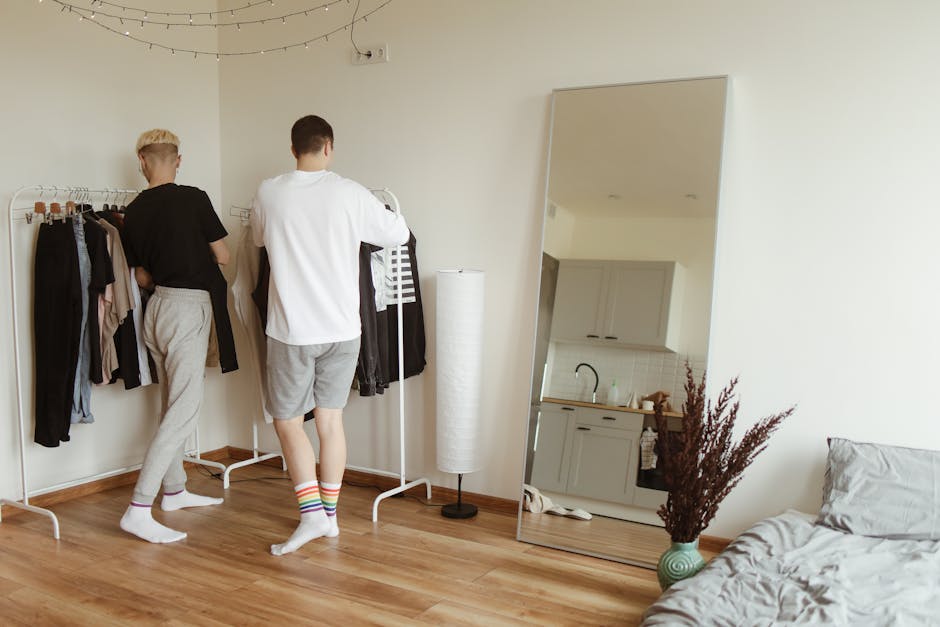 Two men standing in a bedroom, organizing clothes on a clothing rack, reflecting in a large mirror.