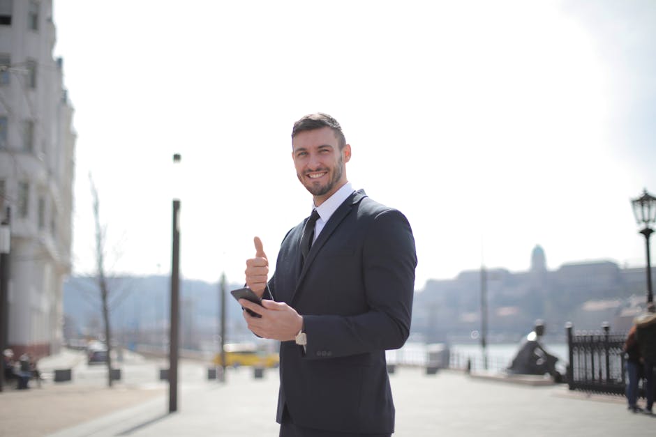Smiling businessman in a suit giving thumbs up and holding a smartphone outdoors in a city setting.