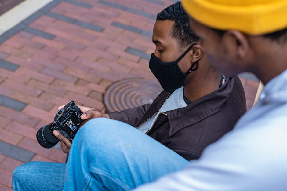A photographer in a face mask reviewing shots on a DSLR camera while sitting outdoors.