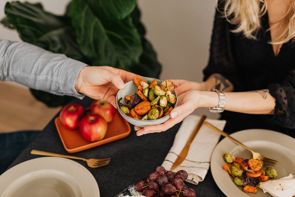 Two hands sharing a bowl of colorful roast vegetables on a table with plates, fruits, and cutlery.