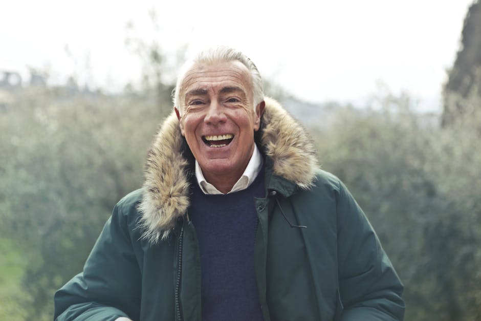 Elderly man with a cheerful smile outdoors wearing a fur-trimmed jacket.
