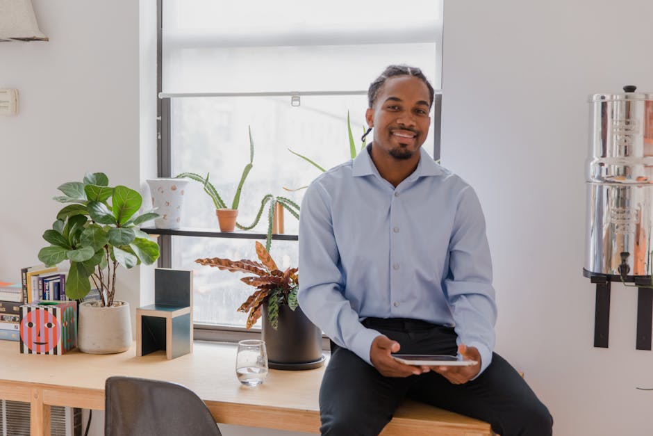 Cheerful man sitting on a desk by the window holding a tablet, surrounded by indoor plants.